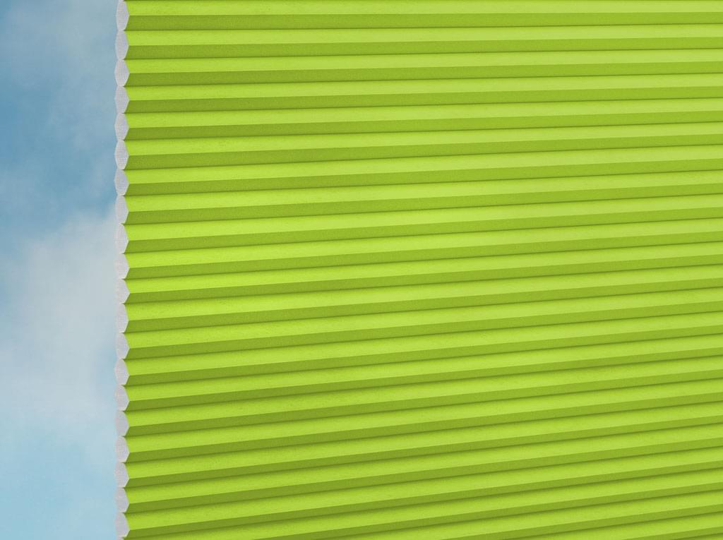Eine Nahaufnahme des DUETTE® Fixé Unix (1.7672) Grün Plissees von Duetteshop, teilweise geöffnet, mit einem sanften blauen Himmel auf der linken Seite.