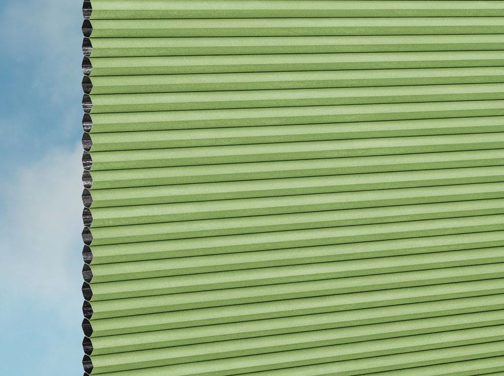 Eine Nahaufnahme des DUETTE® Fixé Unix Dark (3.7471) Grün Plissee nach Maß von Duetteshop, mit einer hellgrünen Wabenstruktur auf der rechten Seite und einem himmelblauen Hintergrund auf der linken Seite.
