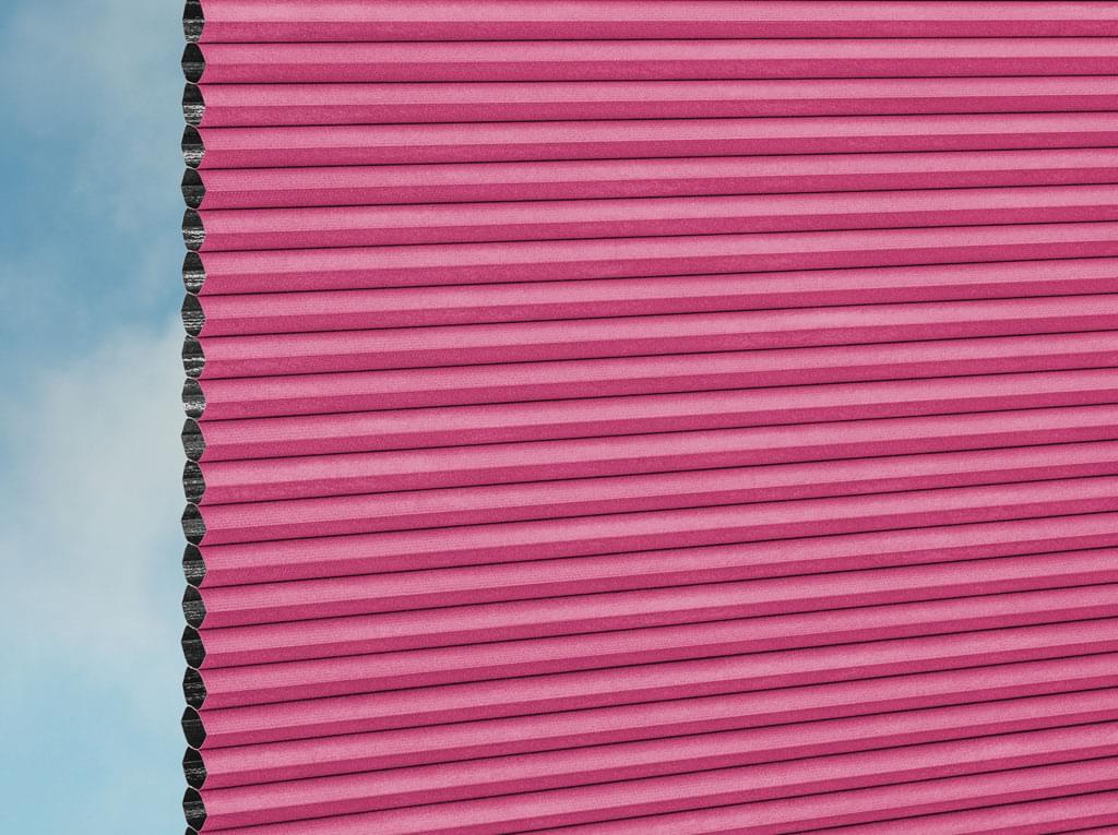 Eine Nahaufnahme der DUETTE® Fixé Unix Dark (3.7493) Rosa Fensterjalousien von Duetteshop zeigt die rechte Seite in Rosa, wobei die linke Kante eine Wabenstruktur aufweist und im Hintergrund ein blauer Himmel mit Wolken zu sehen ist.