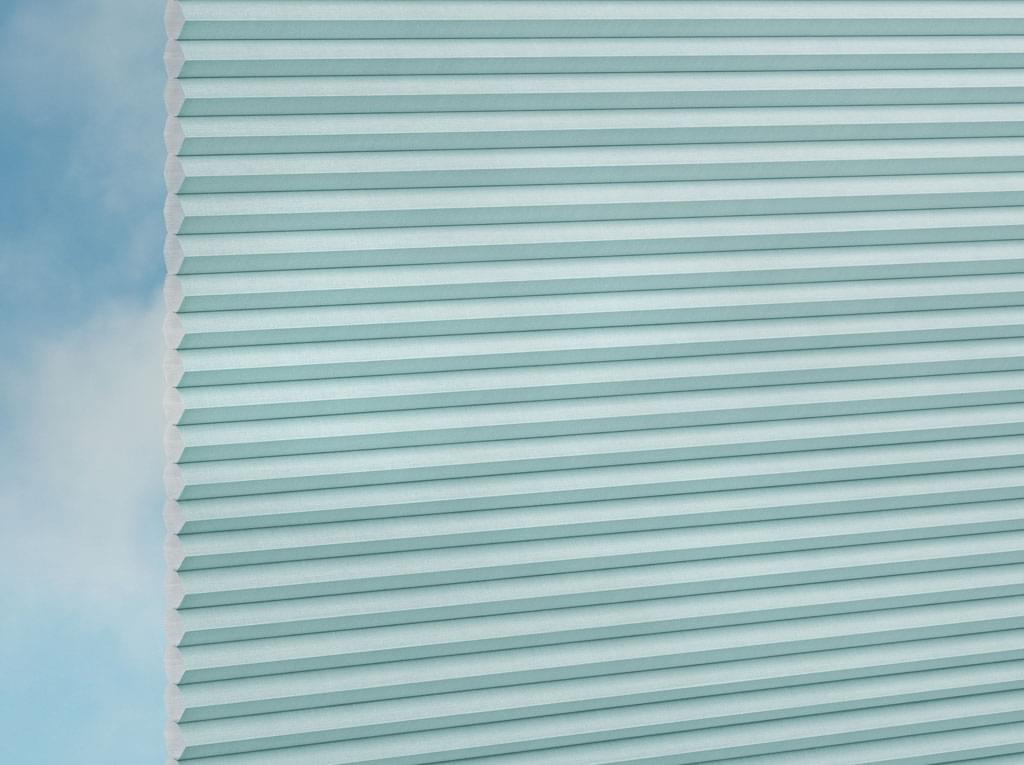 Detailansicht der DUETTE® Fixé Elan (2.7703) Türkis Wabenrollos von Duetteshop mit hellblauem Plisseestoff, teilweise vor einem sanft verschwommenen blauen und weißen Hintergrund dargestellt.