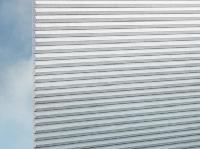 Eine Nahaufnahme von DUETTE® Gradient (3.0802) Türkis von Duetteshop zeigt die weiße, plisseeähnliche, geriffelte Oberfläche mit horizontalen Rillen vor einem blauen Himmel mit weichen Wolken im Hintergrund.