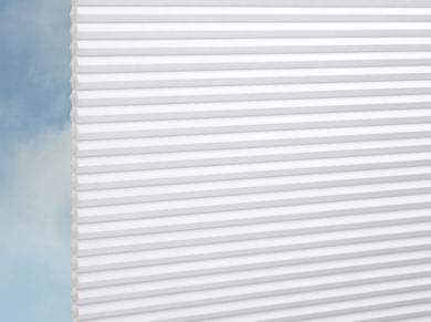 Nahaufnahme von DUETTE® Architella® Elan (3.7001) Weiß von Duetteshop, weiße Zellulose-Jalousien mit Wabenstruktur, teilweise heruntergezogen vor einem blauen Himmel mit leichten Wolken.