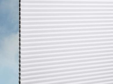 Nahaufnahme der weißen DUETTE® Architella® Elan Dark (4.6601) Weiß Wabenrollos von Duetteshop, Plissee, die ein Fenster mit blauem Himmel und leichten Wolken im Hintergrund teilweise verdecken.