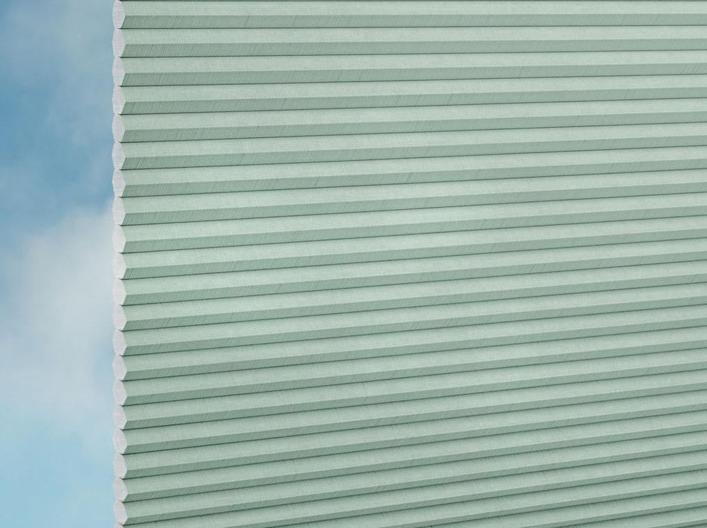 Nahaufnahme der DUETTE® Fixé Batiste (4.6073) Grün Zellulosetöne von Duetteshop, teilweise ausgezogen vor einem unscharfen blau-weißen Himmelshintergrund. Die sechseckige Zellstruktur ist entlang der linken Kante sichtbar.