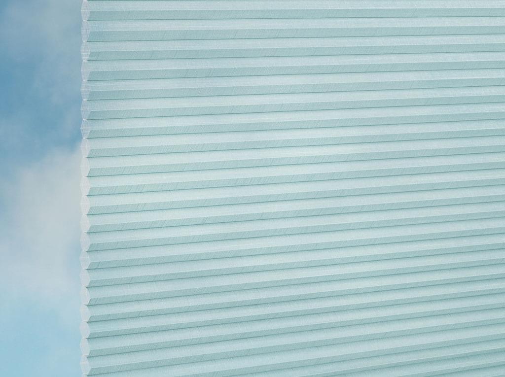 Eine Nahaufnahme von DUETTE® Fixé Batiste Sheer (4.9349) Türkis von Duetteshop in Hellblau, vor einem sanft verschwommenen, vom Himmel inspirierten Hintergrund.