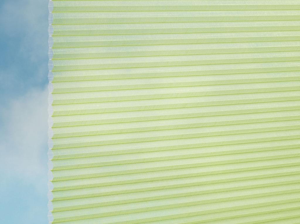 Eine Nahaufnahme des DUETTE® Fixé Batiste Sheer (4.9375) Grün von Duetteshop, die die blassgrünen Falten und die Wabenstruktur zeigt, die das Licht sanft filtern, während links der blaue Himmel zu sehen ist.