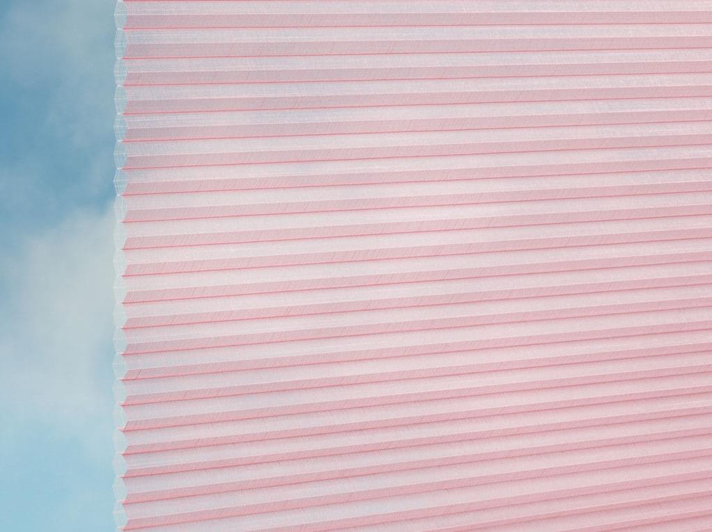 Nahaufnahme des DUETTE® Fixé Batiste Sheer (4.9397) Rosa von Duetteshop, ein hellrosa Plissee mit Wabenstruktur, das einen weichen, wolkenblauen Himmel im Hintergrund teilweise verdeckt.
