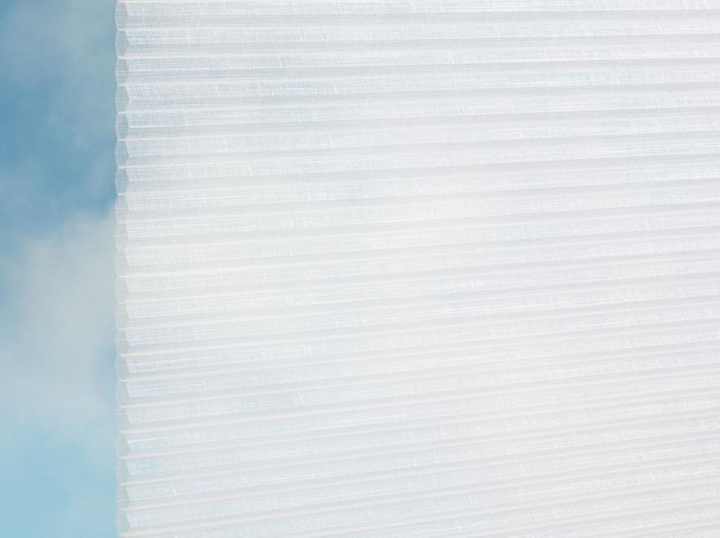 DUETTE® Fixé Batiste Sheer (4.6502) Weiß von Duetteshop besteht aus weißen, lichtdurchlässigen Wabenschirmen, die bei teilweiser Öffnung weiches natürliches Licht filtern. Ein blauer Himmel mit leichten Wolken im Hintergrund unterstreicht die luftige Atmosphäre.